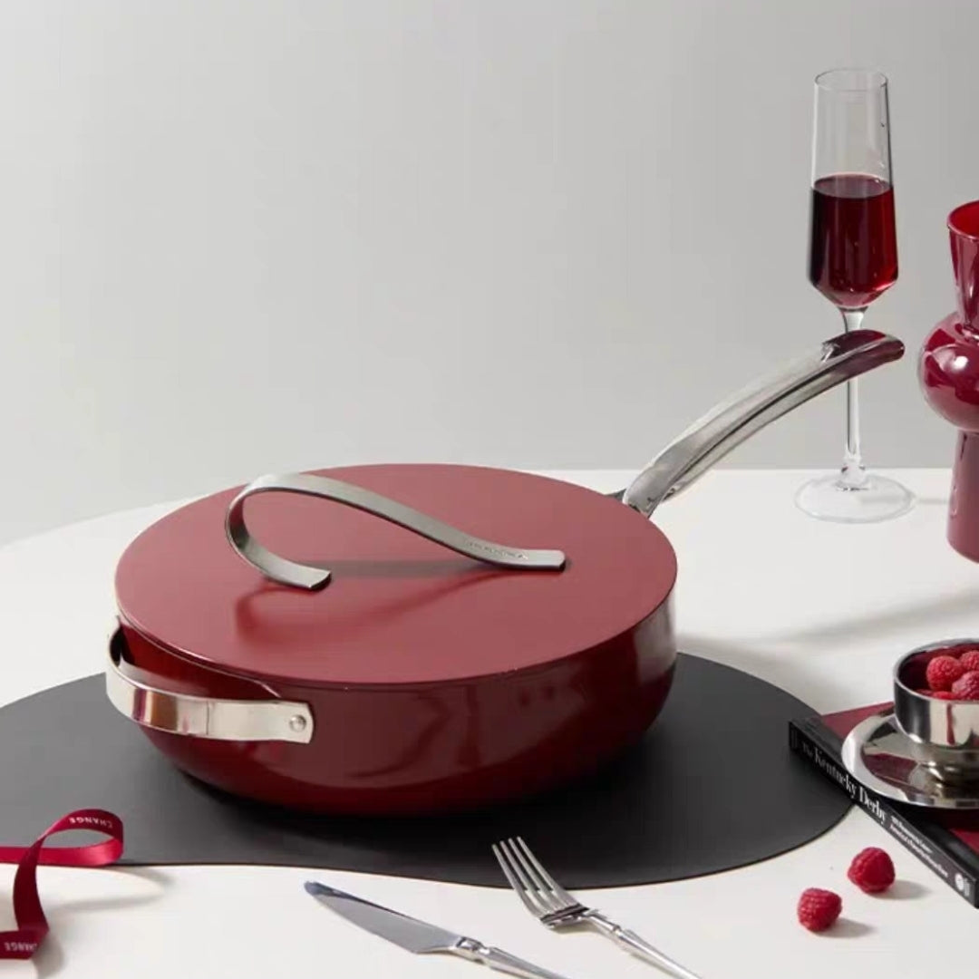 Red non-stick ceramic frying pan with silver handle on a white surface with wine and raspberries.