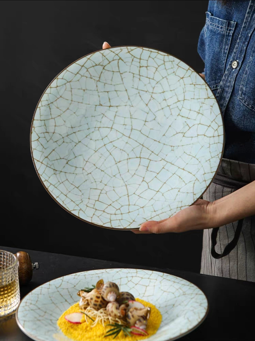 Person holding a ceramic plate with a cracked glaze design, showcasing a dish of food on a dark background.