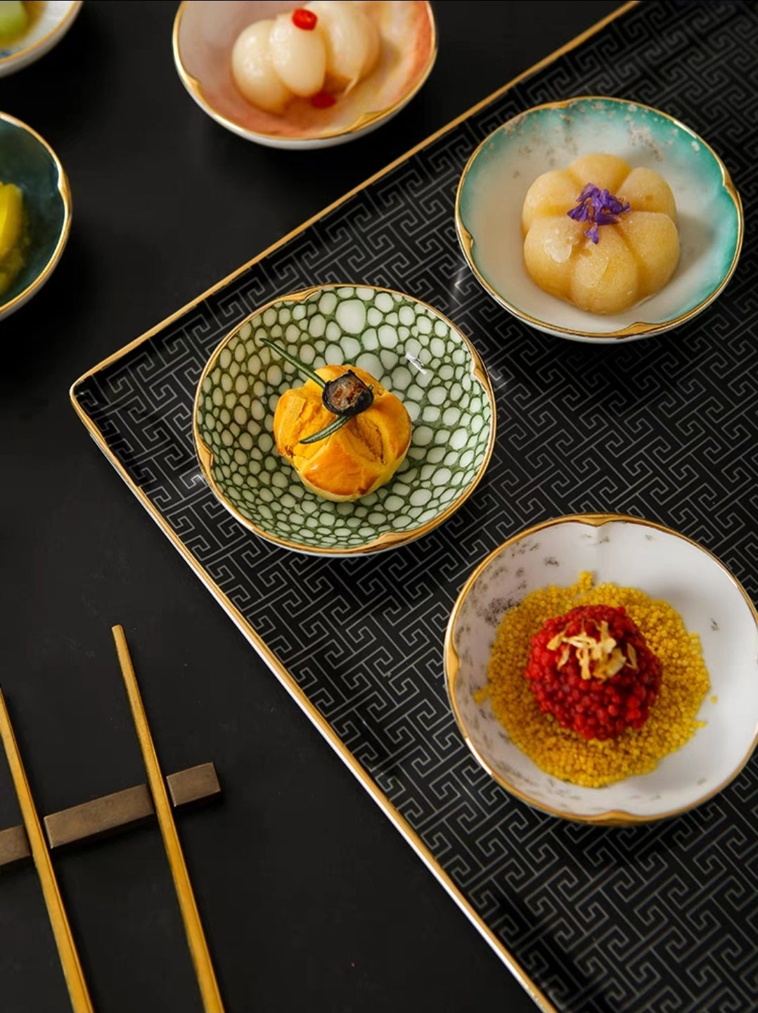 Assorted ceramic dipping bowls with golden-trimmed edges, displayed with wooden chopsticks on textured serving tray.
